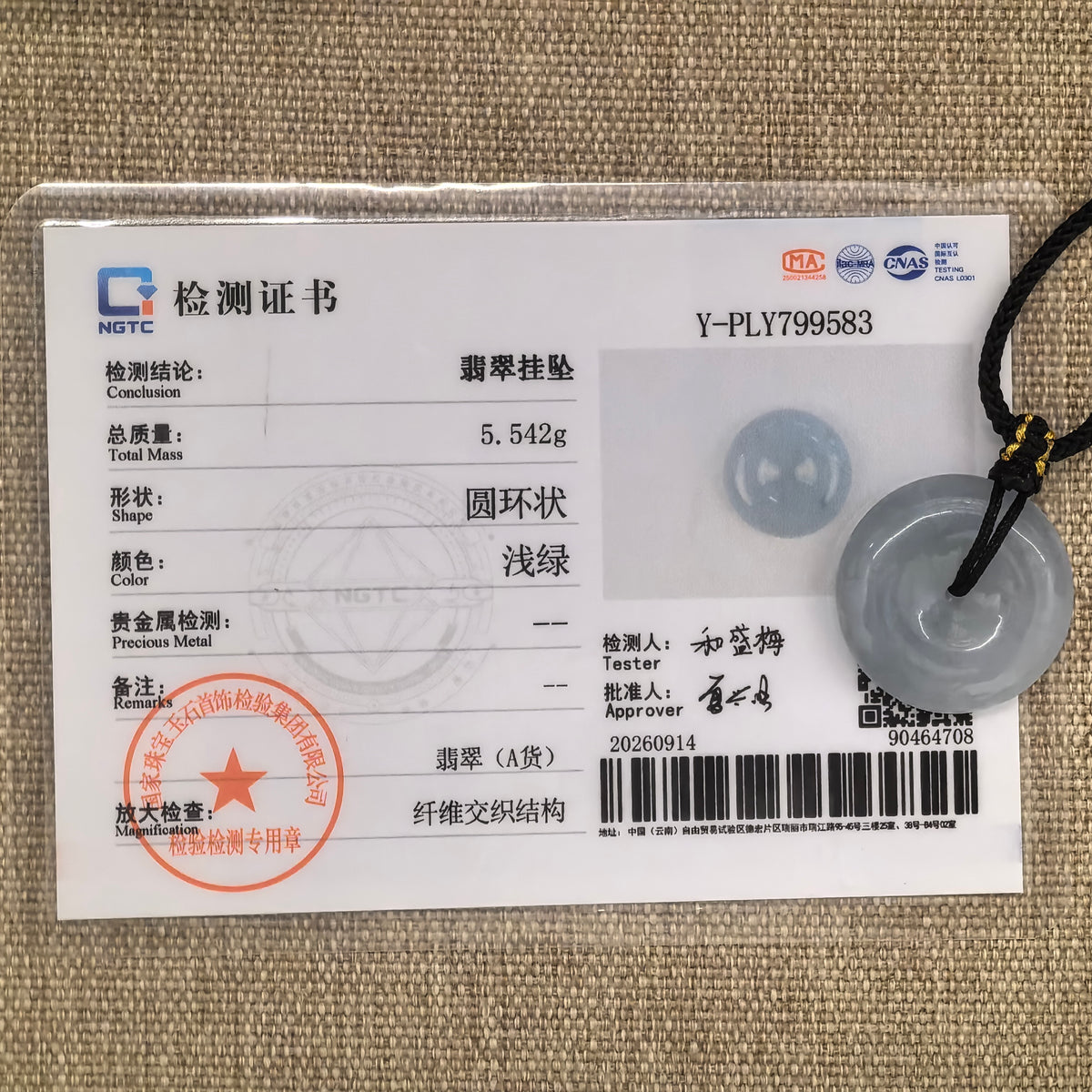 Pale blue jadeite Peace Pendant with its black cord beside the matching NGTC certificate on tan woven fabric.