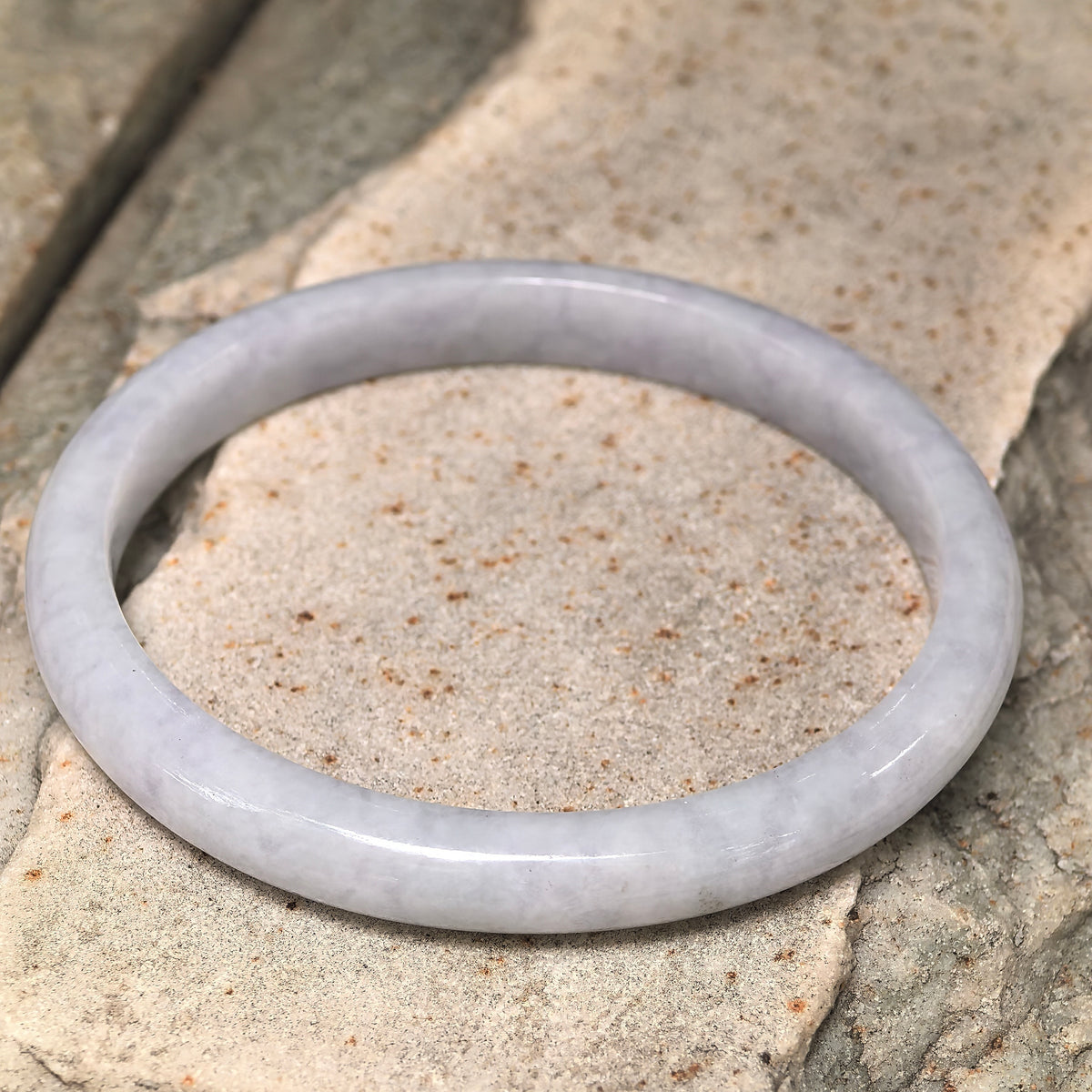 Angled view of a white jadeite D-shape bangle resting on stone, showing its rounded outer profile and flat inner surface.