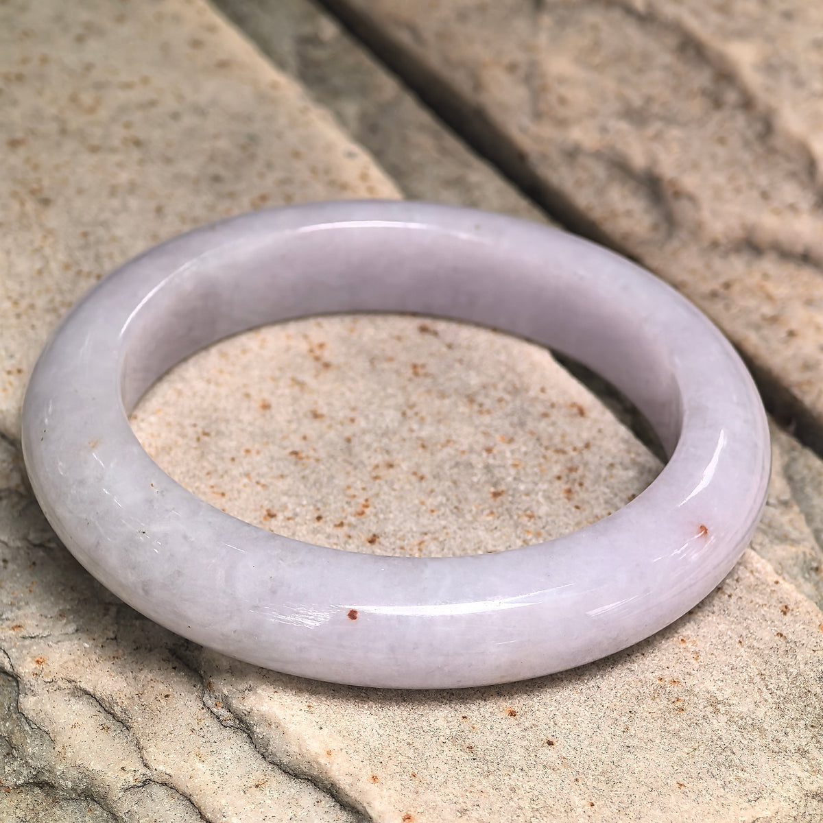 Angled view of a pale lavender jadeite D-shape bangle on stone, showing golden-brown flecks, a flat inner surface and rounded outer profile.