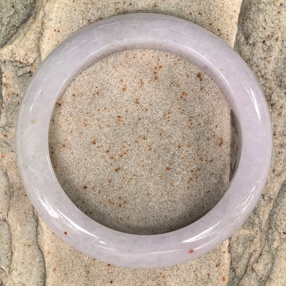 Overhead view of a pale lavender jadeite bangle on textured stone, showing its circular outline and scattered golden-brown flecks.