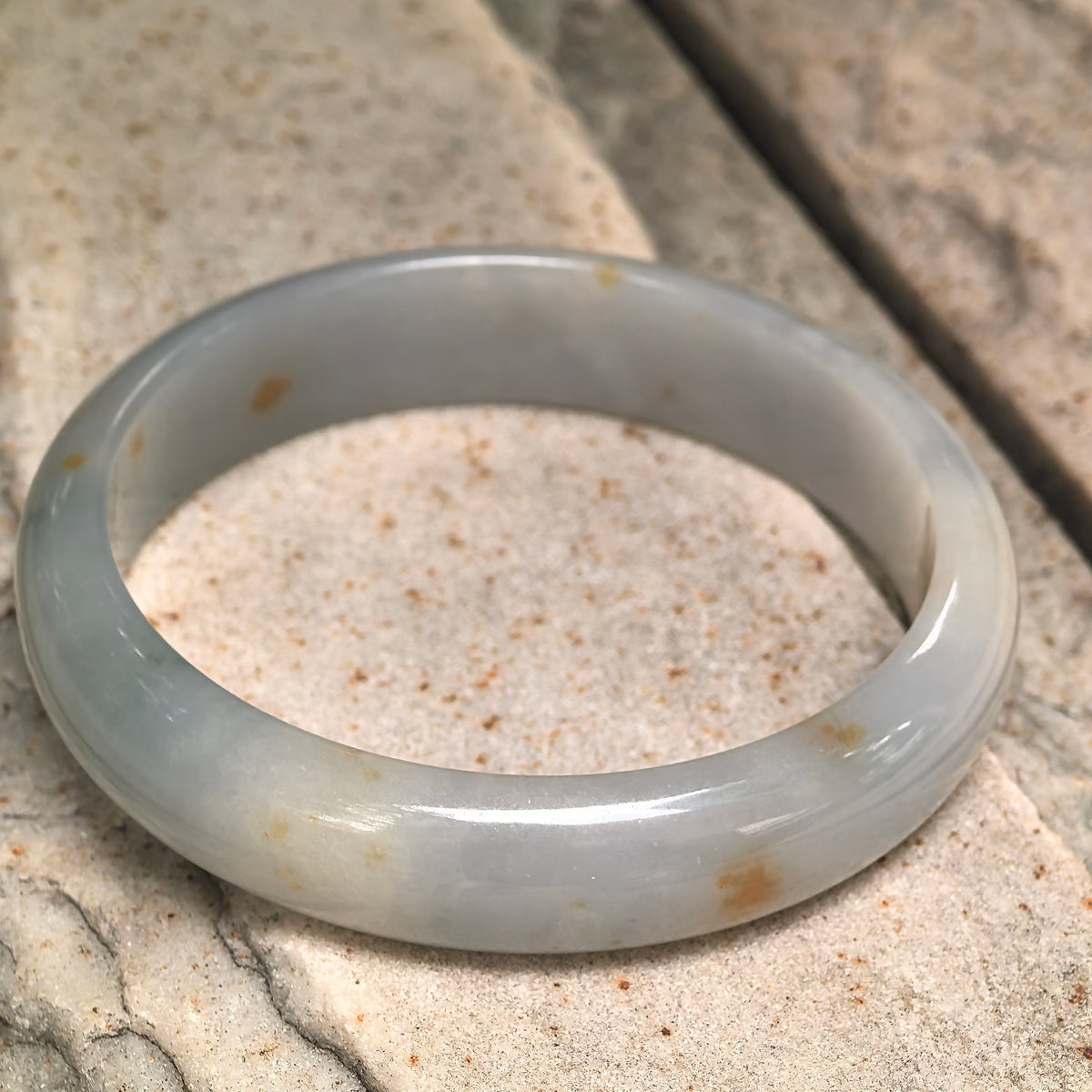 White and pale green jadeite bangle resting at an angle on stone, showing golden-brown flecks, a flat inner surface and rounded outer profile.