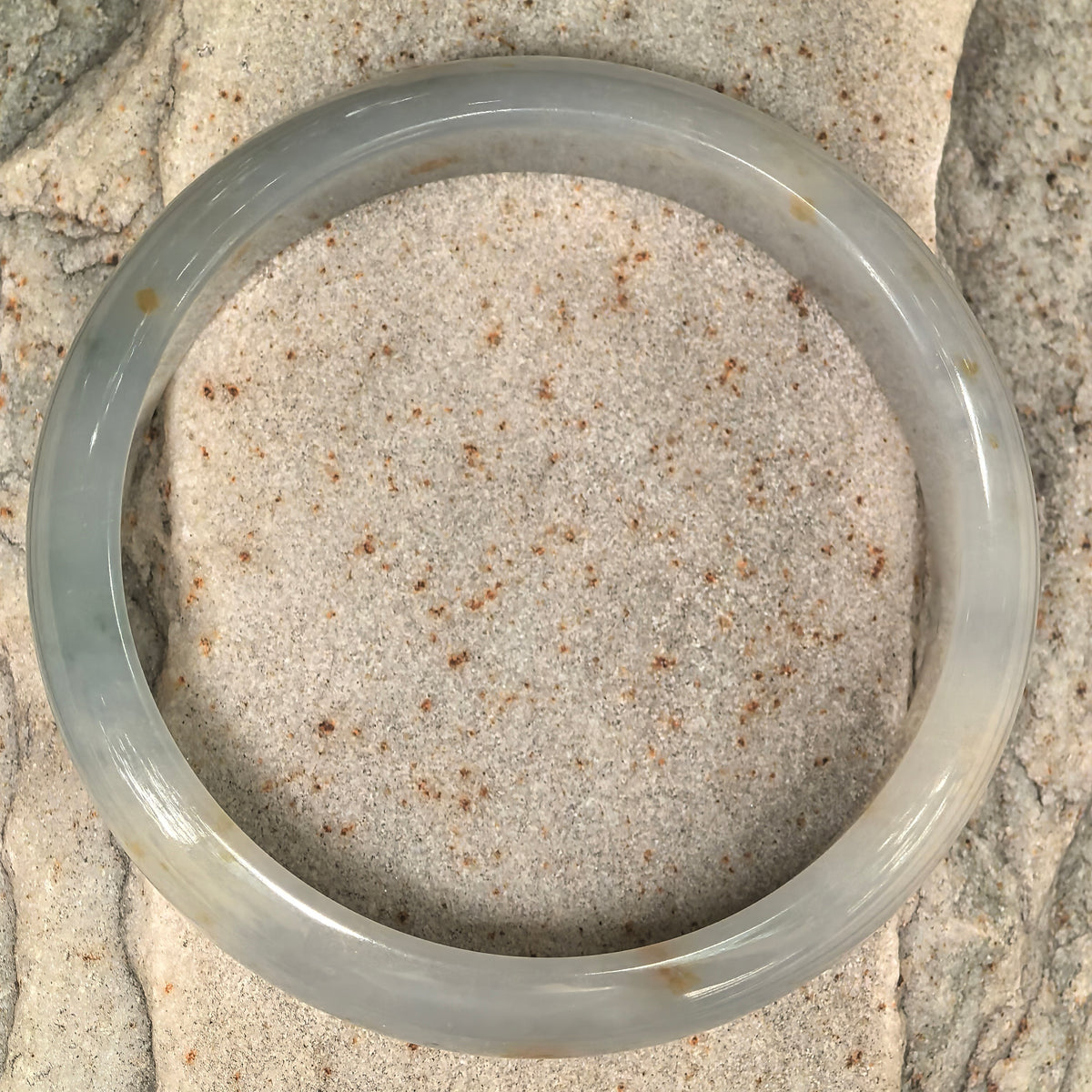 Overhead view of a white and pale green jadeite D-shape bangle on textured stone, showing the distribution of natural golden-brown flecks.