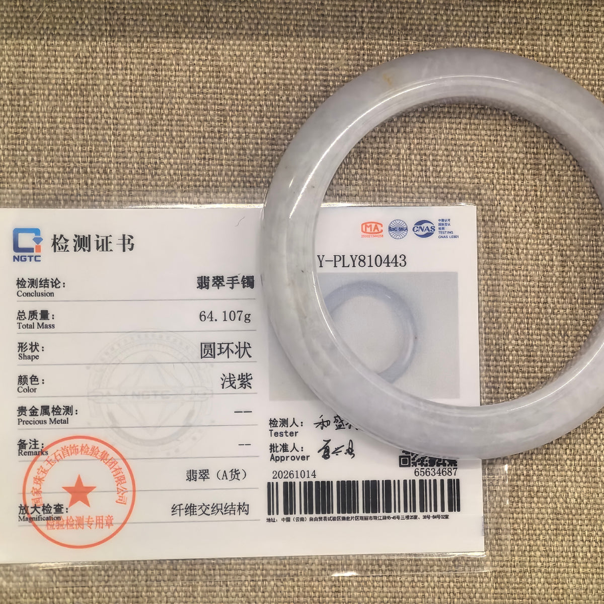 Pale lavender jadeite bangle beside its matching NGTC certificate on tan woven fabric.