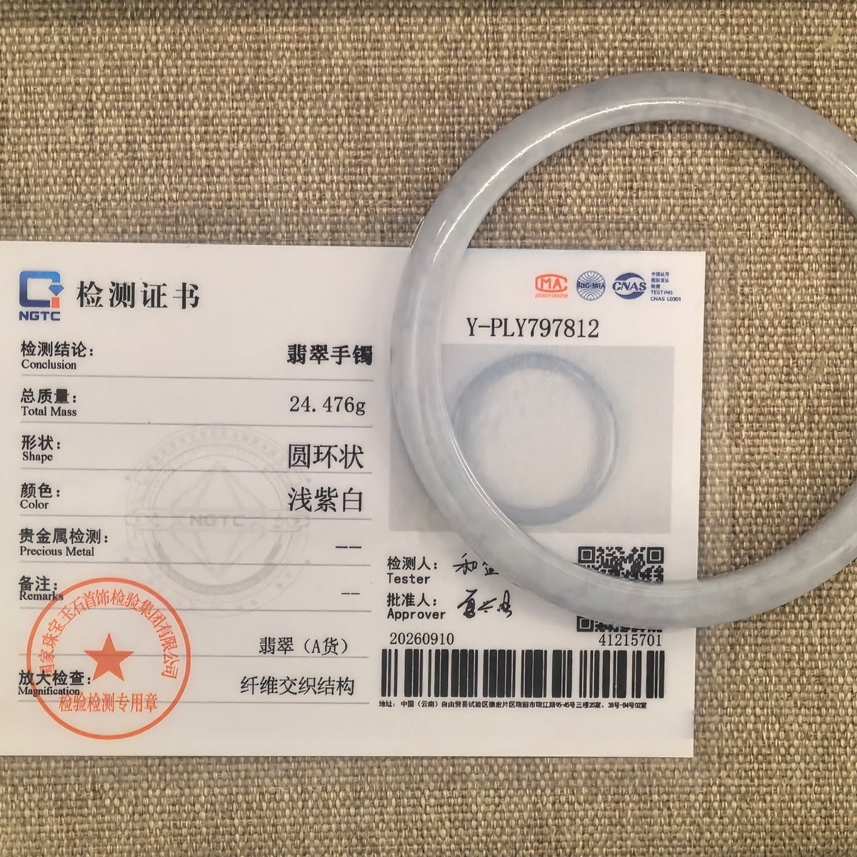 Slim white jadeite bangle displayed with its matching NGTC certificate on woven fabric.