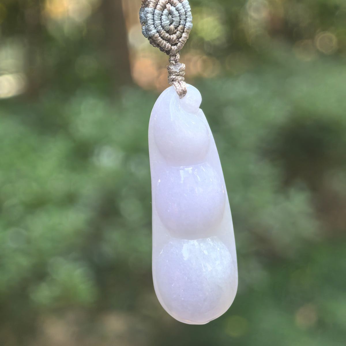 Pale lavender jadeite Bean Pod Pendant hanging from a braided cord outdoors, showing three rounded beans and a curled tip.