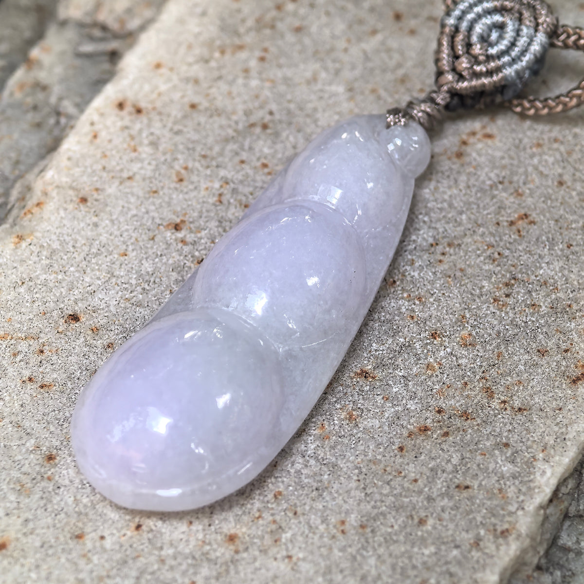 Angled close-up of a lavender jadeite Bean Pod Pendant on stone, showing its raised bean carving and rounded contours.
