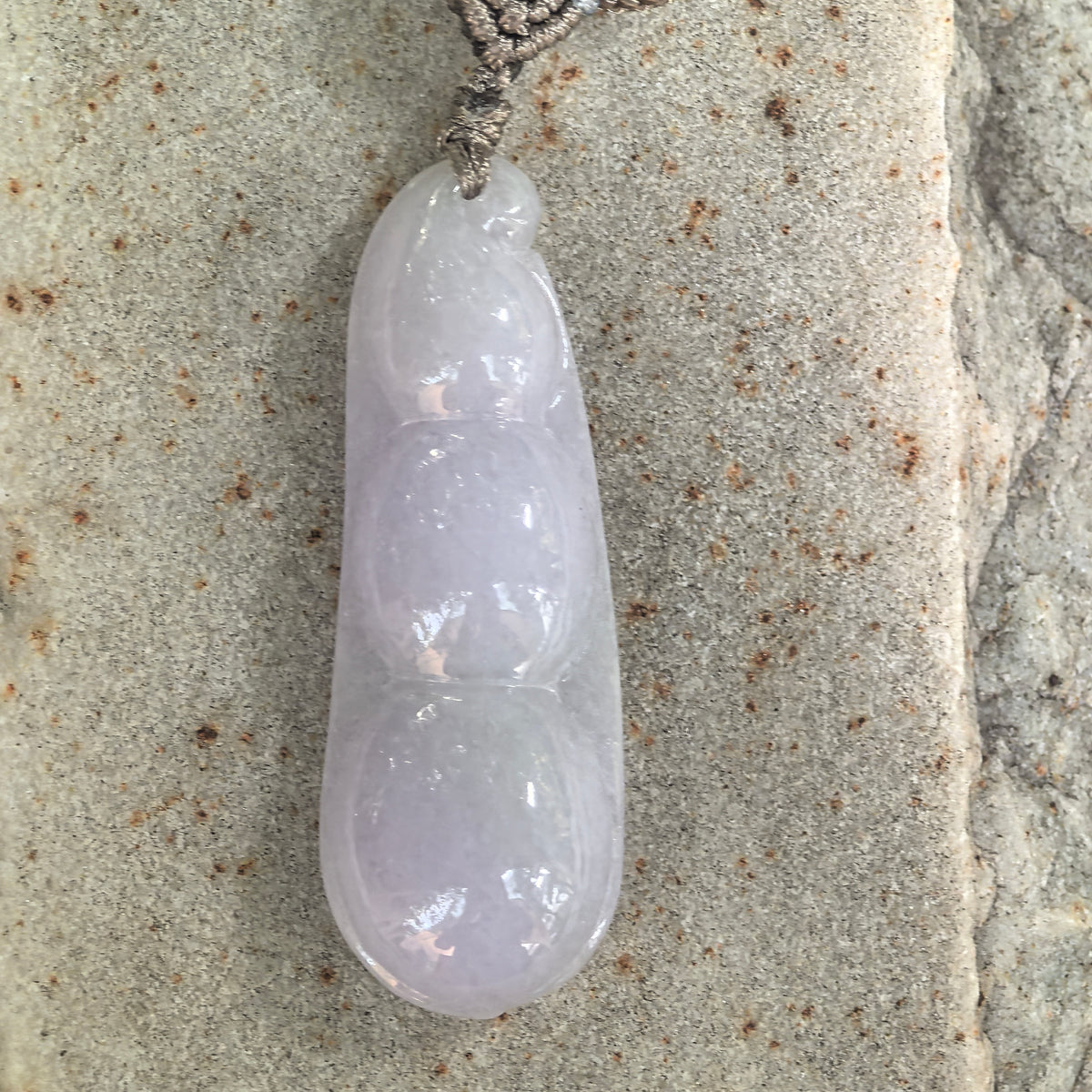 Overhead view of a pale lavender jadeite Bean Pod Pendant lying on stone, showing the full three-bean design and polished surface.