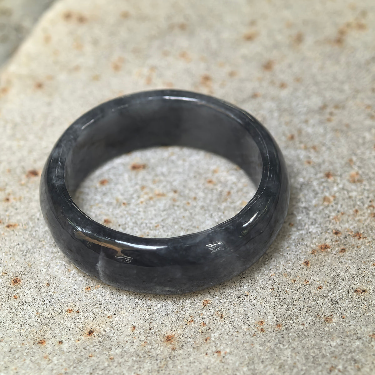 Angled view of a gray-black jadeite ring band resting on stone, showing its rounded outer profile and polished inner surface.
