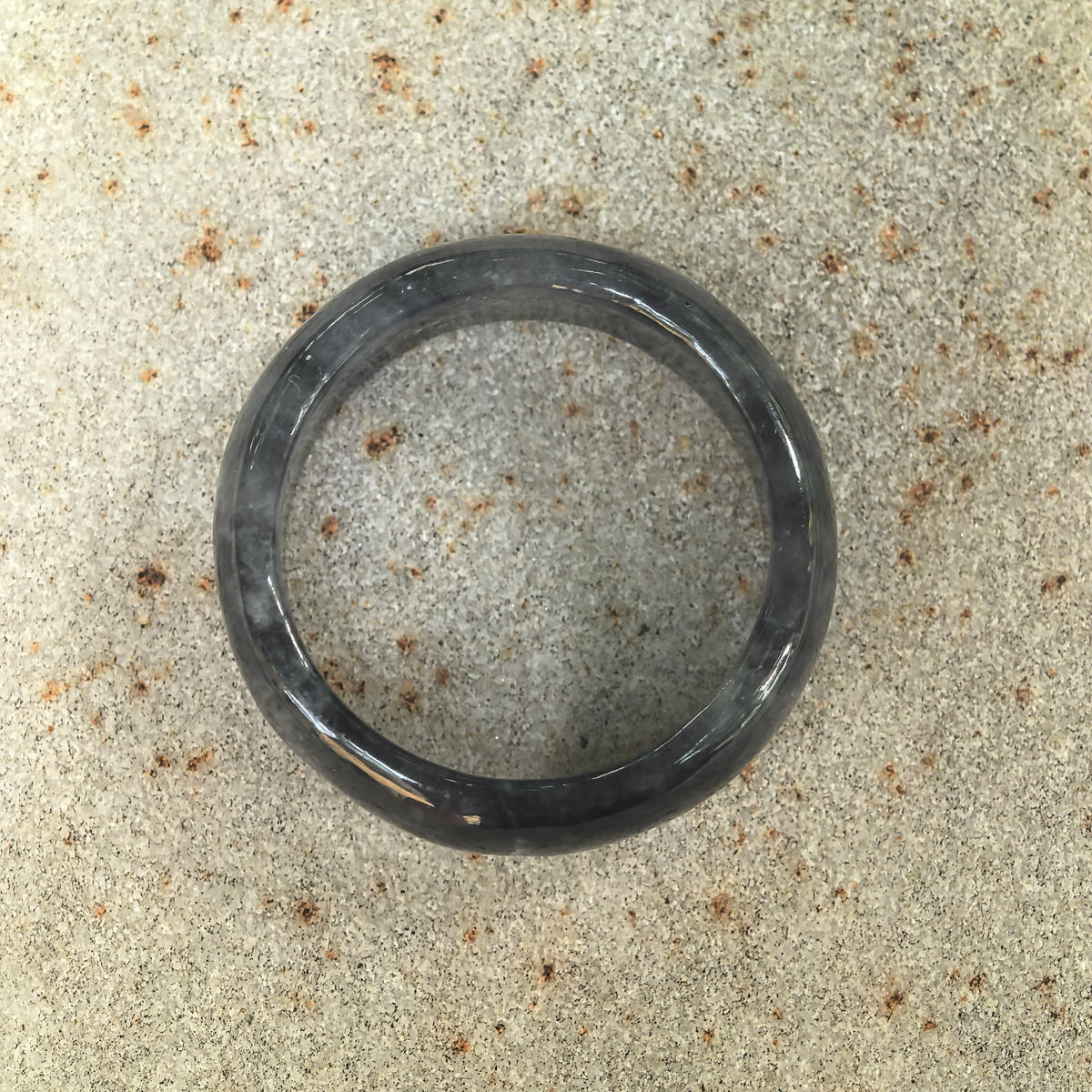 Overhead view of a gray-black jadeite ring band on stone, showing its circular outline and smoky natural patterns.