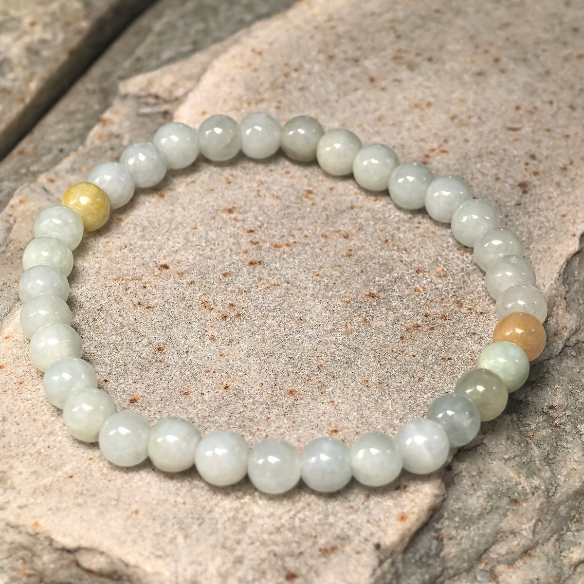 Angled view of the pale green jadeite bead bracelet on stone, showing two yellow accent beads and softly clouded textures.