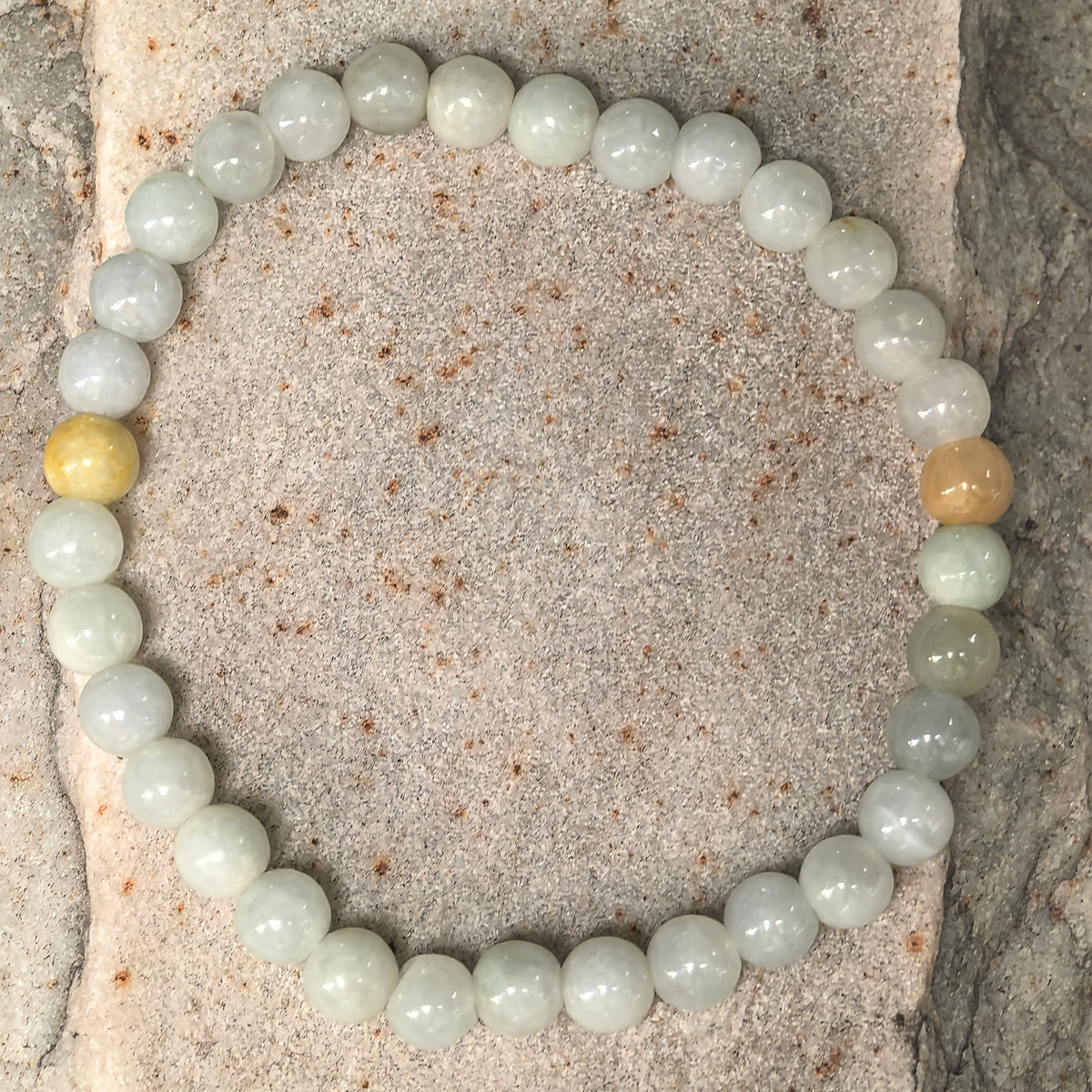 Overhead view of the pale green glutinous jadeite bead bracelet arranged in a circle on stone with two yellow accent beads.
