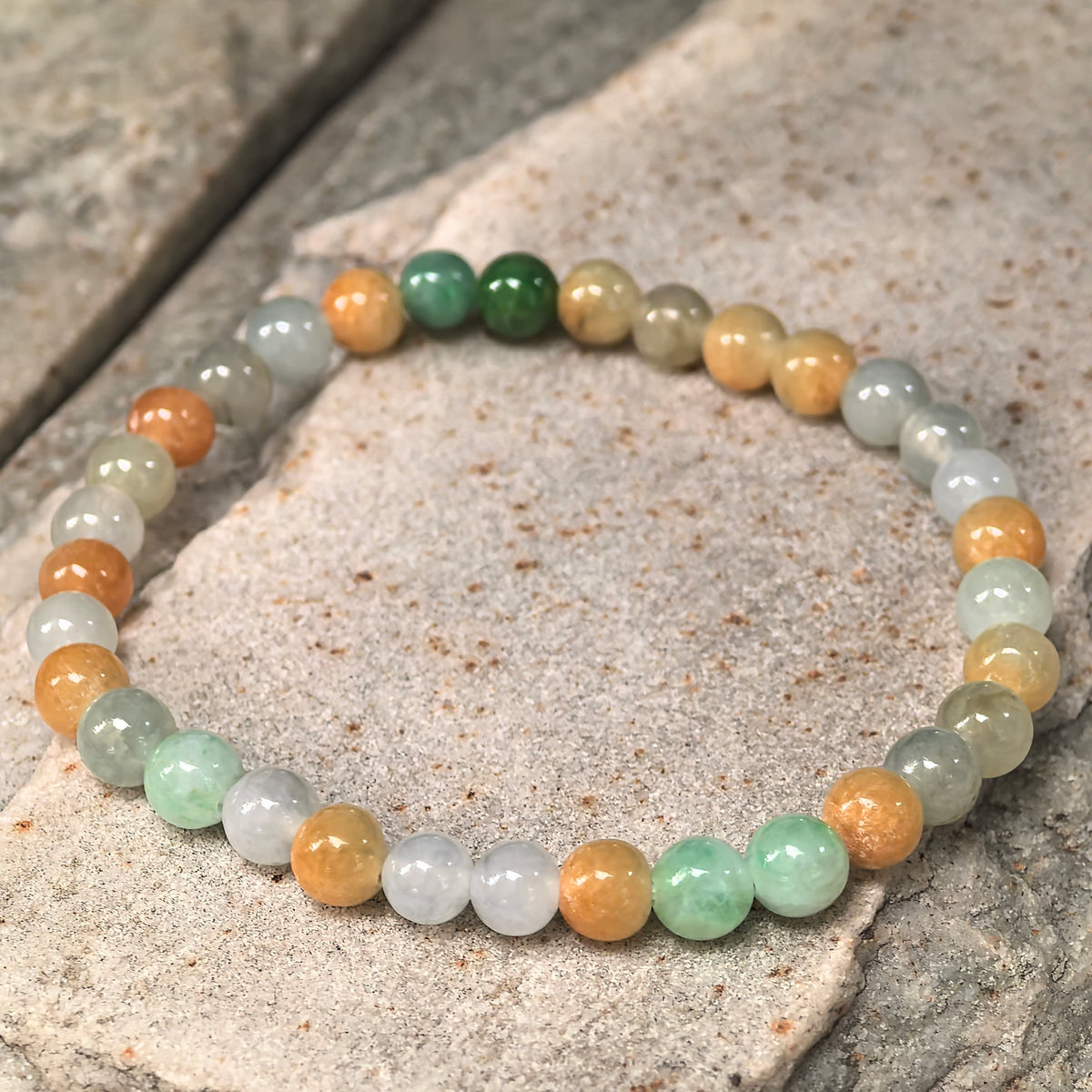Angled view of a multicolor jadeite bead bracelet resting on stone, showing natural variations in bead color and texture.