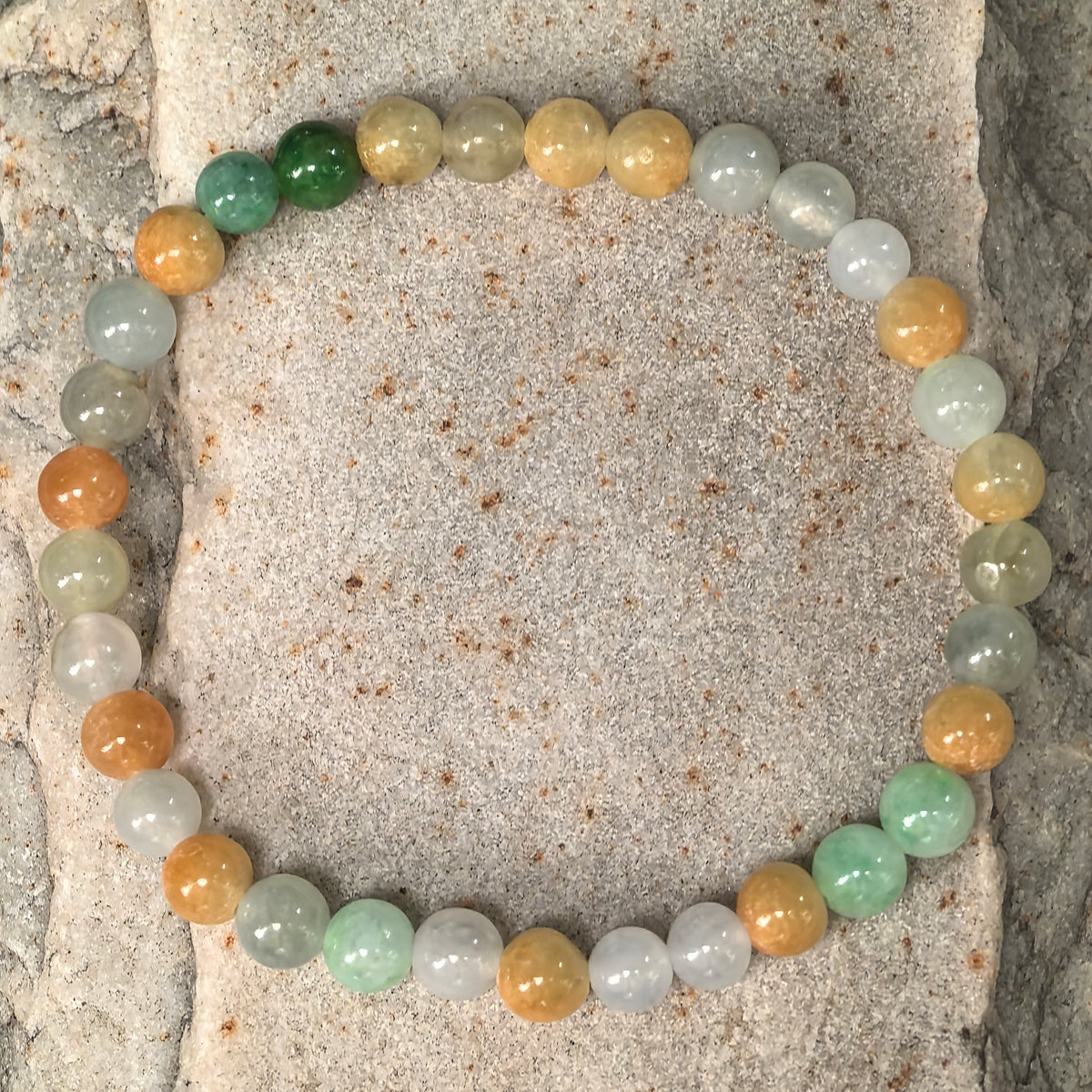 Overhead view of a multicolor jadeite bead bracelet on stone, with warm yellow and orange-brown beads interspersed with green and white beads.