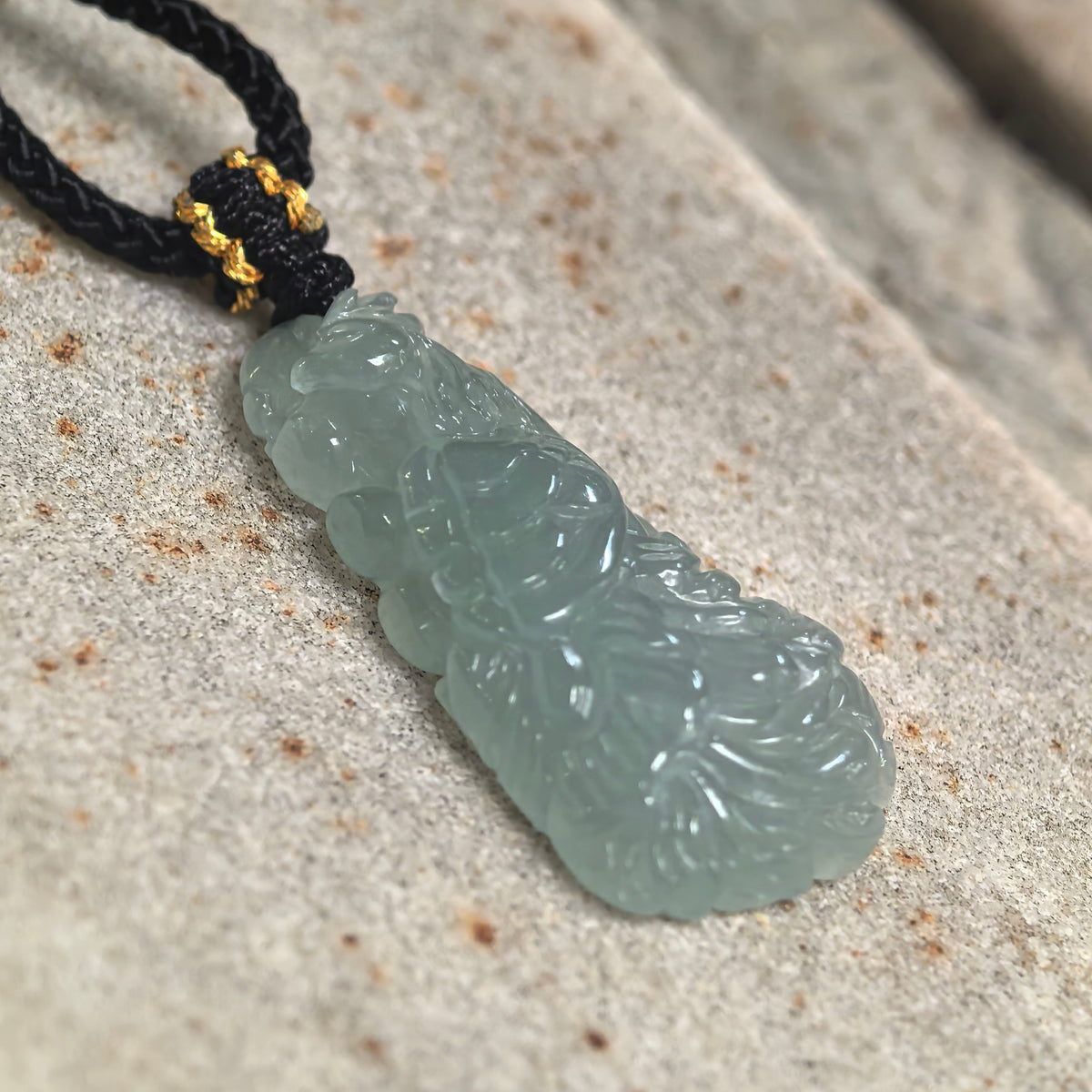 Angled close-up of a light blue jadeite phoenix pendant on stone, showing raised feather details and polished contours.