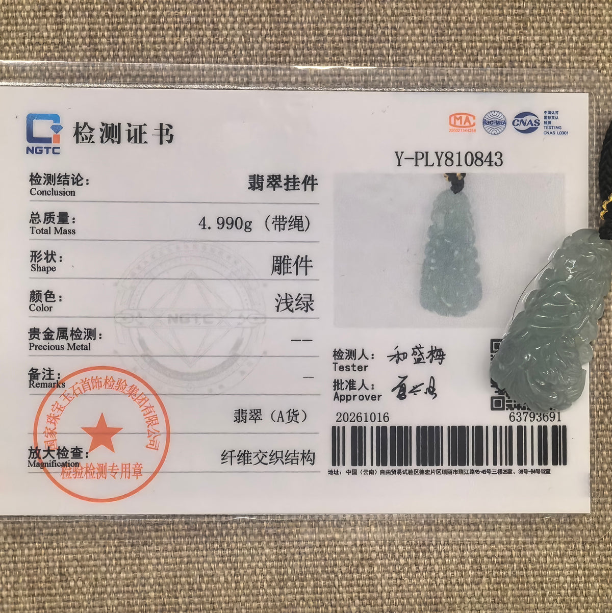 Light blue jadeite phoenix pendant displayed with its matching NGTC certificate on woven fabric.