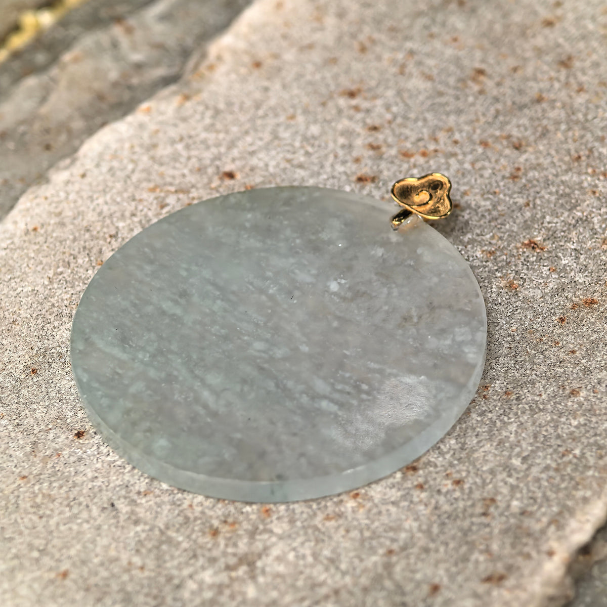 Angled view of the round jadeite Plain Plaque Pendant on stone, showing its polished face, natural white inclusions and decorative gold bail.