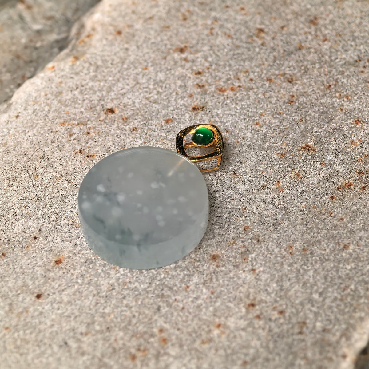Angled view of the round jadeite Plain Plaque Pendant on stone, showing its polished surface, scattered white inclusions and green accent stone in the gold bail.