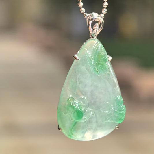 Icy light green jadeite butterfly and lotus pendant hanging from a chain, showing deeper green carvings and an 18K gold bail with diamond accents.