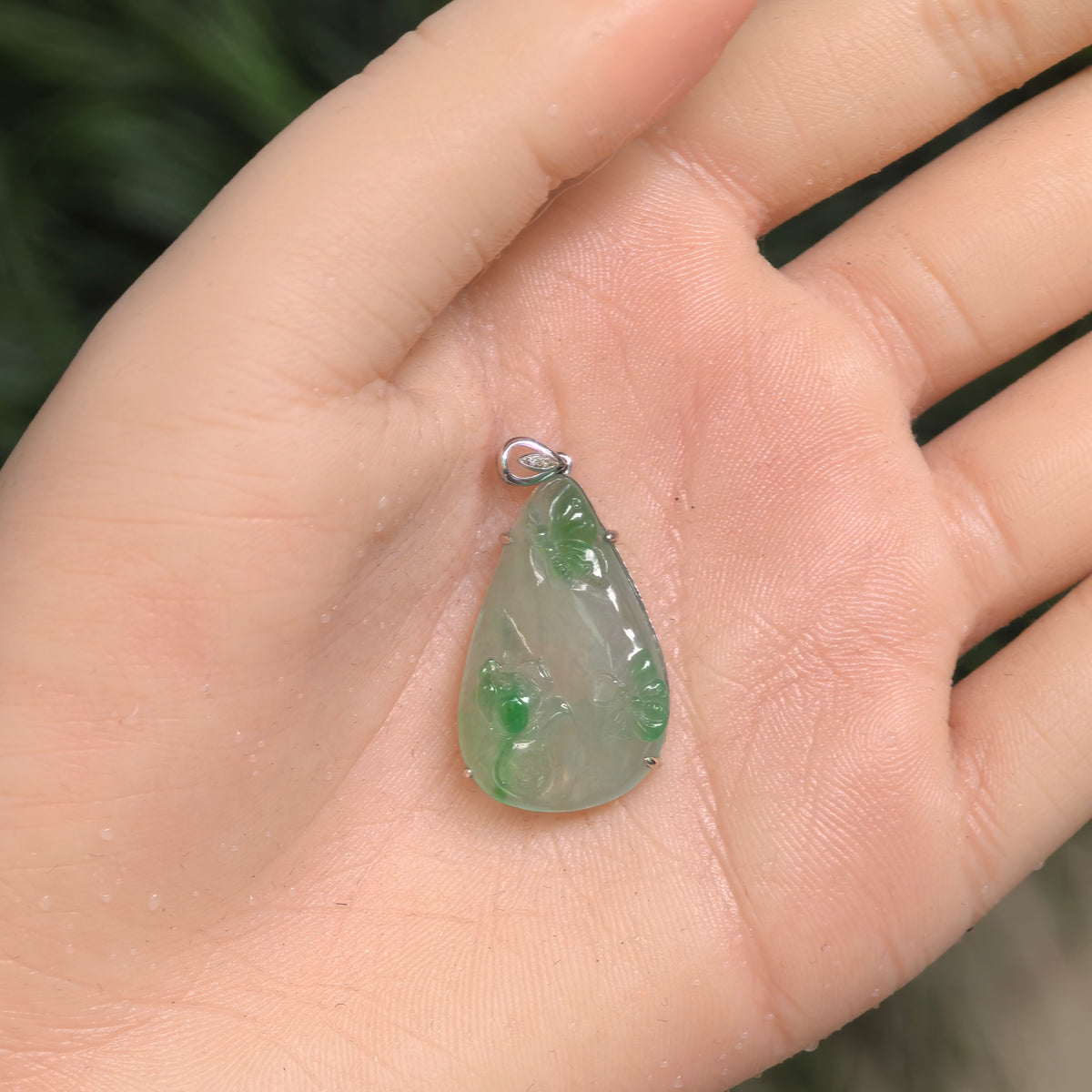 Light green jadeite pendant resting in an open palm, showing its tapered outline, two carved butterflies and a lotus blossom.