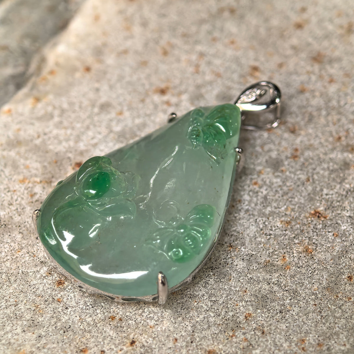 Angled close-up of the jadeite pendant on stone, showing green butterfly wings, raised lotus petals and gold prongs.