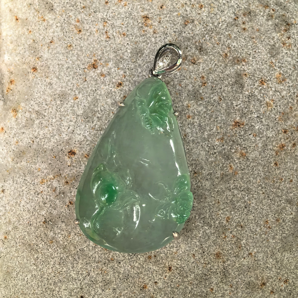 Front view of the light green jadeite pendant on stone, showing two butterflies, a lotus blossom and the curved bail with diamond accents.