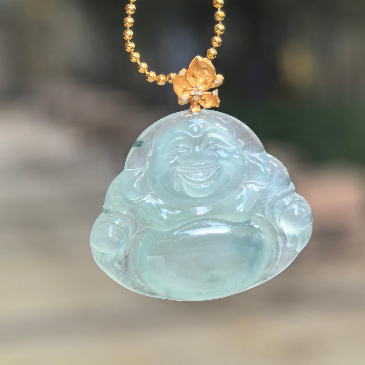 Front view of an icy light green jadeite Laughing Buddha pendant hanging from a chain, showing its smiling face and gold bail with diamond accents.