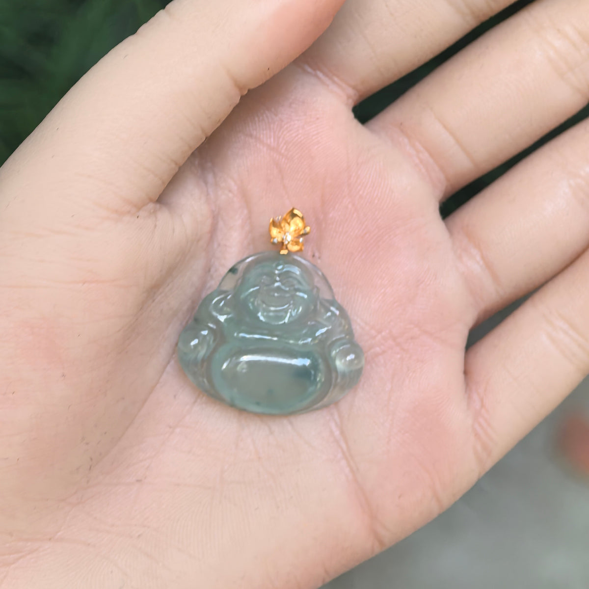 Light green jadeite Laughing Buddha pendant resting in a palm, showing its broad rounded outline and carved belly beneath the gold bail.