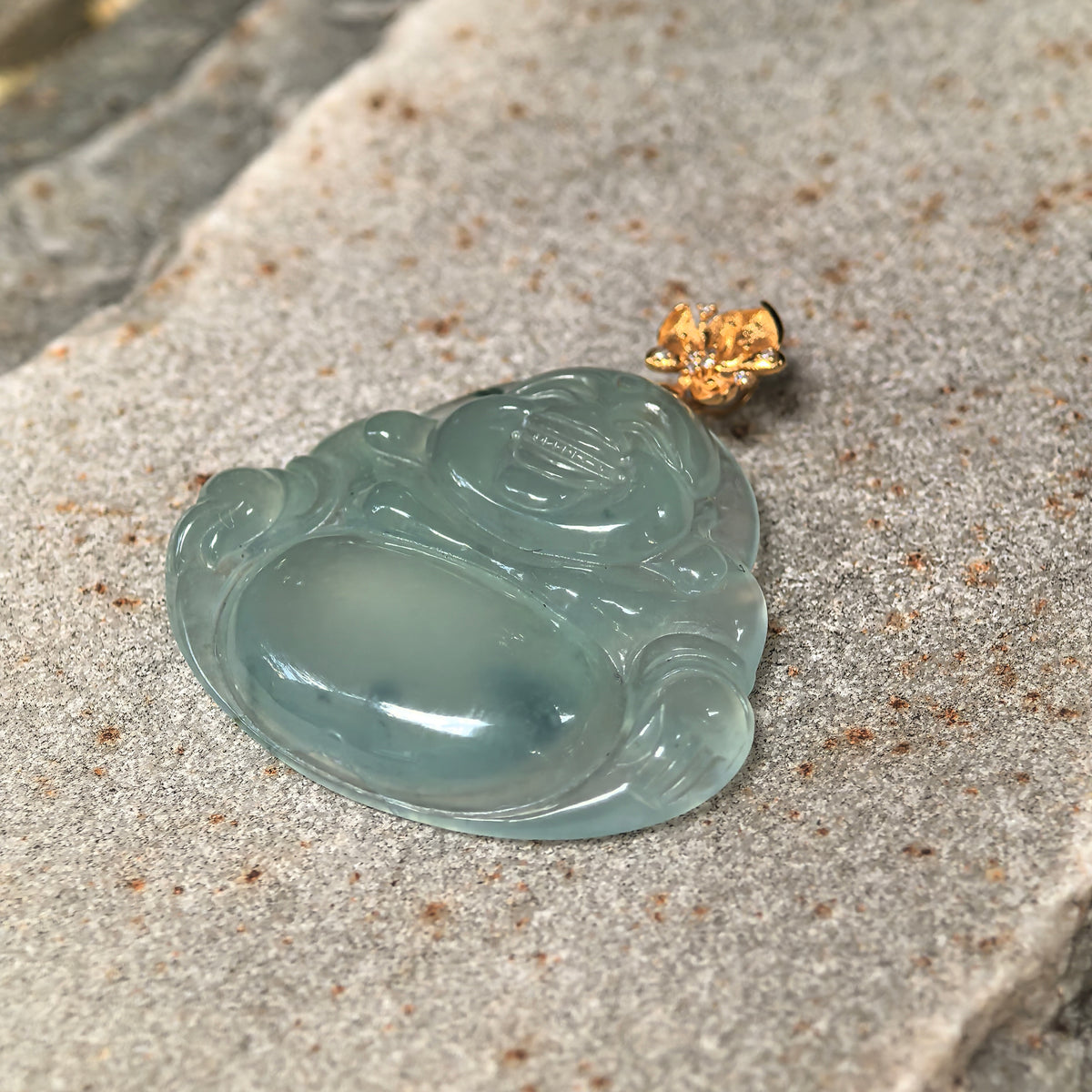 Angled view of a light green jadeite Laughing Buddha pendant on a stone surface, highlighting the relief carving and polished contours.
