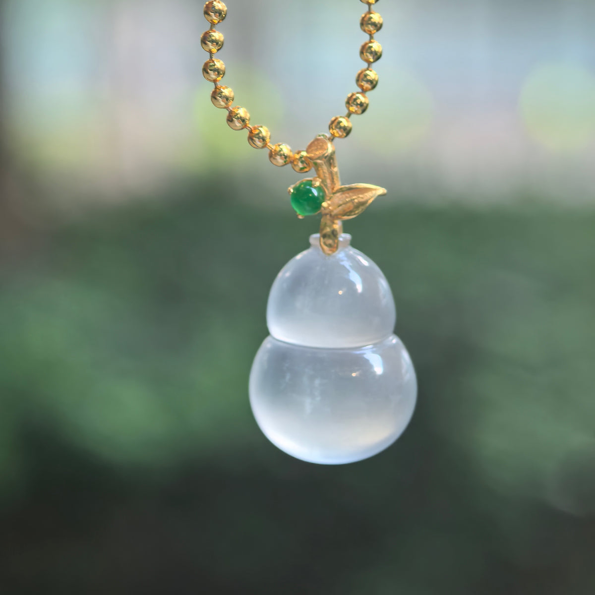 Icy translucent white jadeite Gourd Pendant hanging from a chain, with bright highlights, an 18K gold leaf bail, and a small green accent stone.