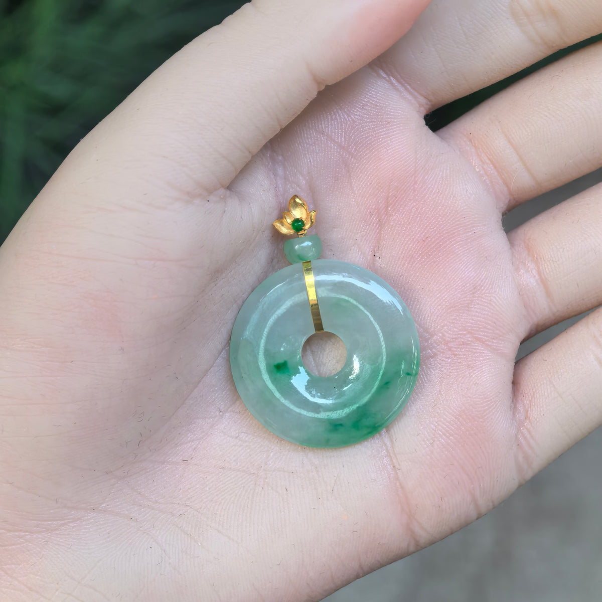 Light green jadeite Peace Pendant resting in an open palm, showing floating green patterns, a central opening, and an 18K gold floral bail.