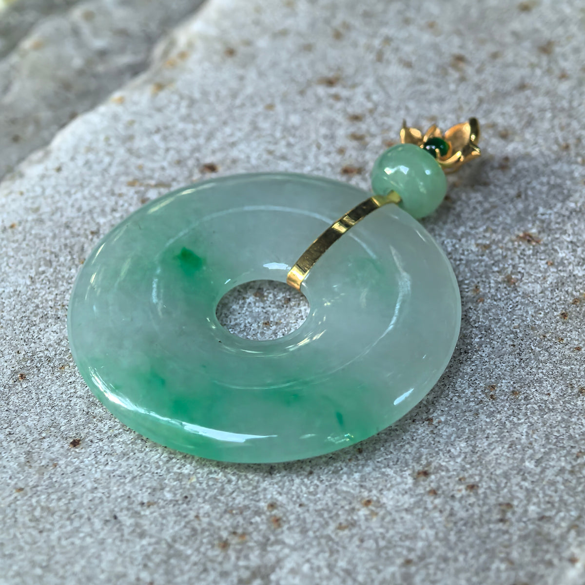 Angled close-up of the light green jadeite Peace Pendant on stone, showing its curved surface, brighter green patches, and narrow gold strip.