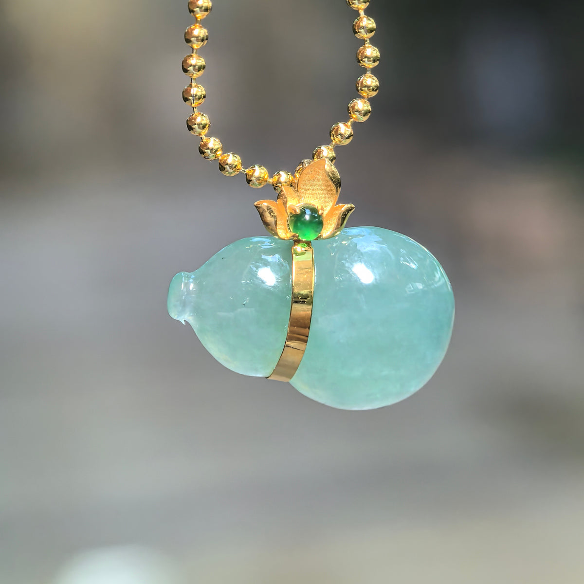 Icy green jadeite horizontal Gourd Pendant hanging from a chain, showing its rounded lobes, 18K gold waist band and floral bail with a green accent stone.