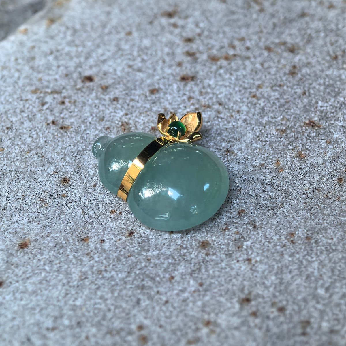 Angled view of the green jadeite Gourd Pendant on stone, showing the rounded carving, gold band around the waist and floral bail.