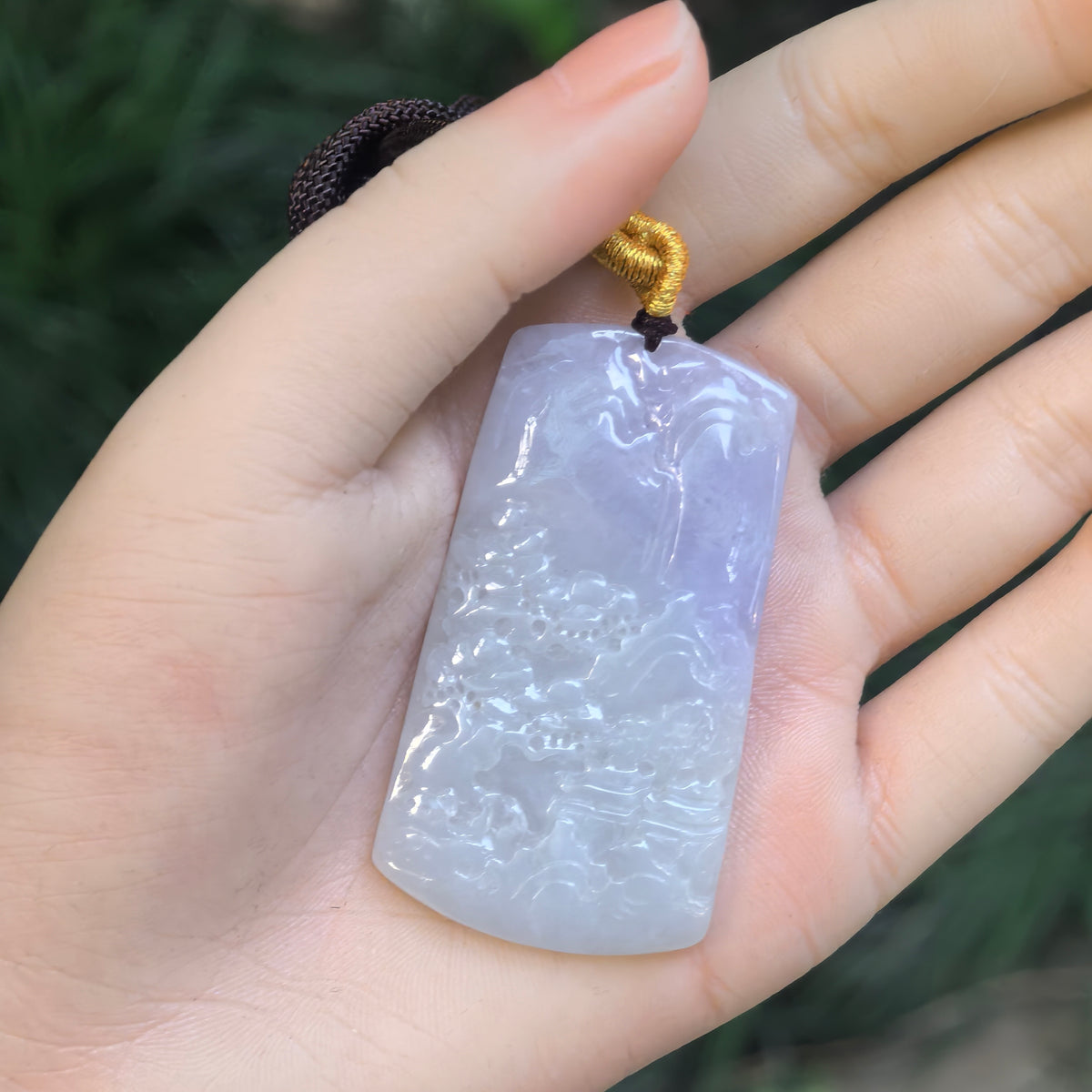 Jadeite landscape plaque pendant resting in a palm, showing its purple section, rectangular shape and carved mountain scenery.