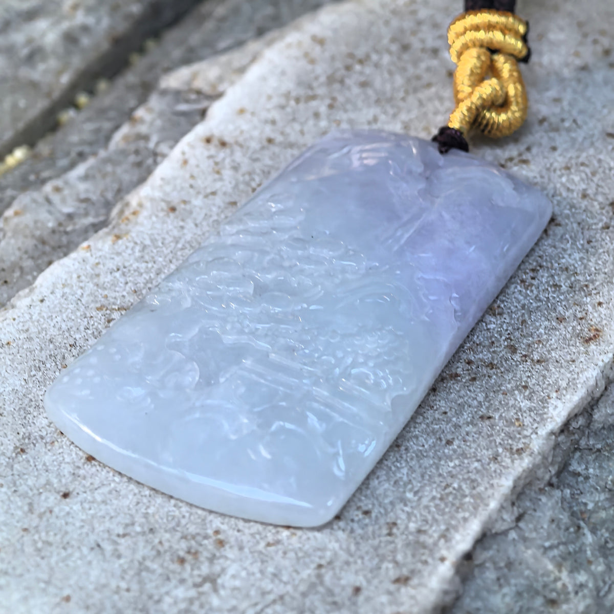 Angled view of a partly purple jadeite landscape plaque pendant on stone, highlighting the relief carving and rounded edges.