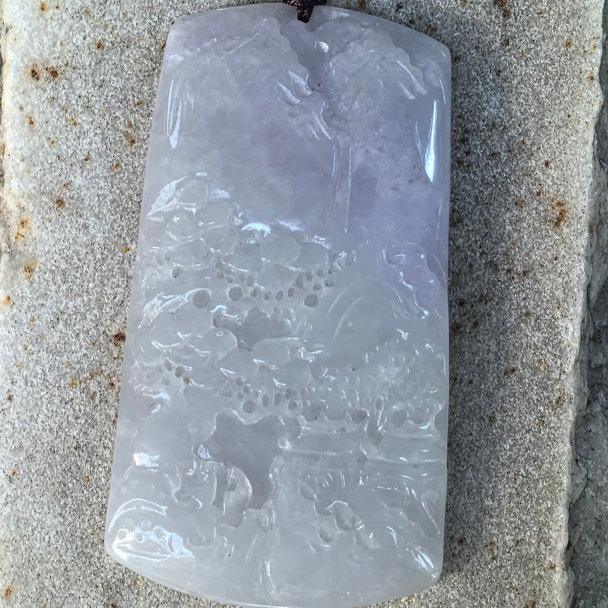 Front close-up of a jadeite landscape plaque pendant on stone, showing mountain ridges, clustered trees and layered rock formations.