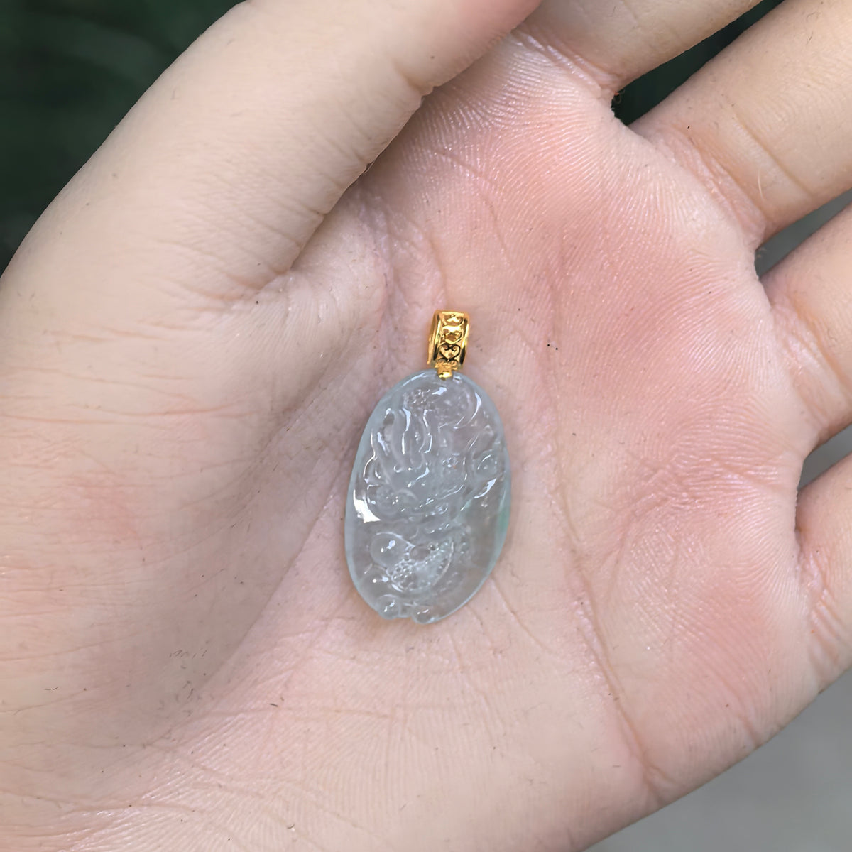 White jadeite dragon pendant resting in a palm, showing its compact oval shape, coiled dragon carving and openwork gold bail.