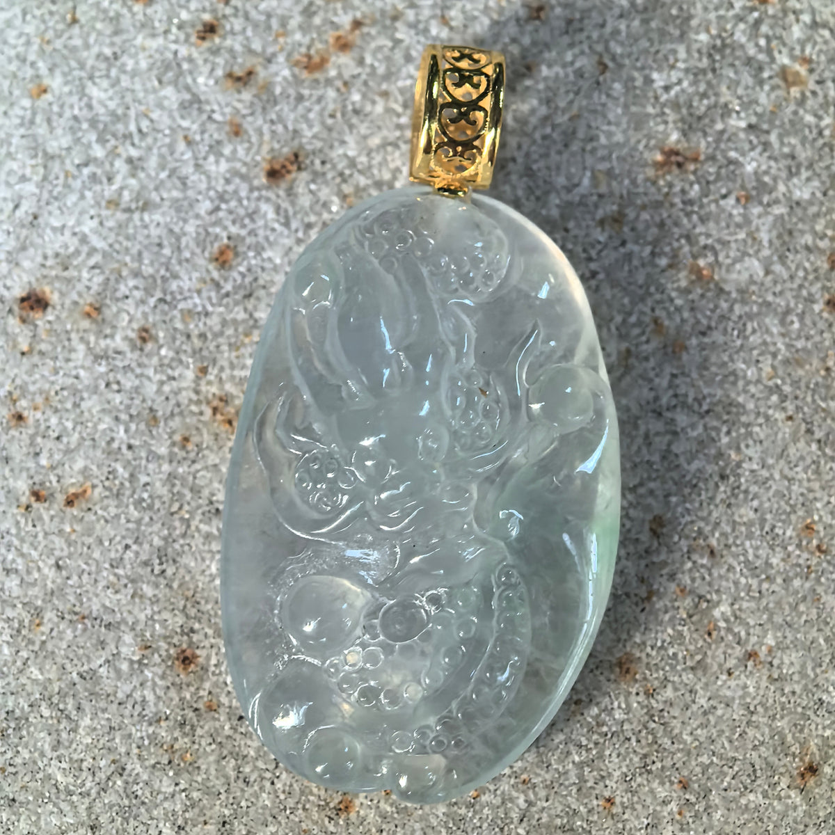 Front close-up of a white jadeite dragon pendant on a stone surface, showing the detailed relief carving and decorative openwork gold bail.