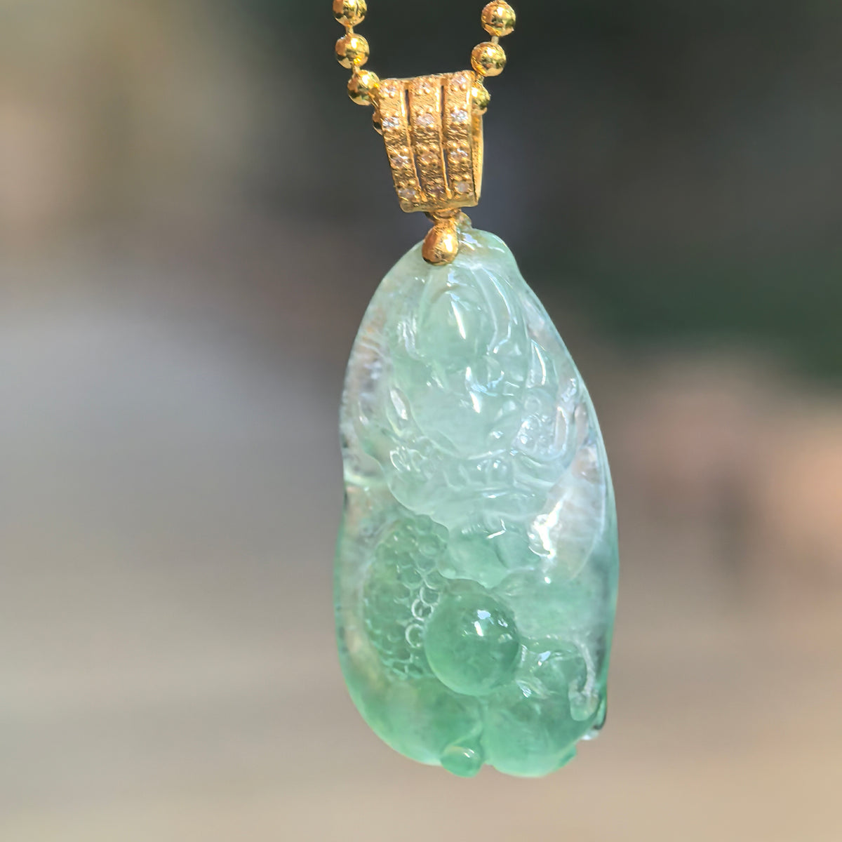 Icy light green jadeite dragon pendant hanging from a chain, showing its relief carving and 18K gold bail with diamond accents.
