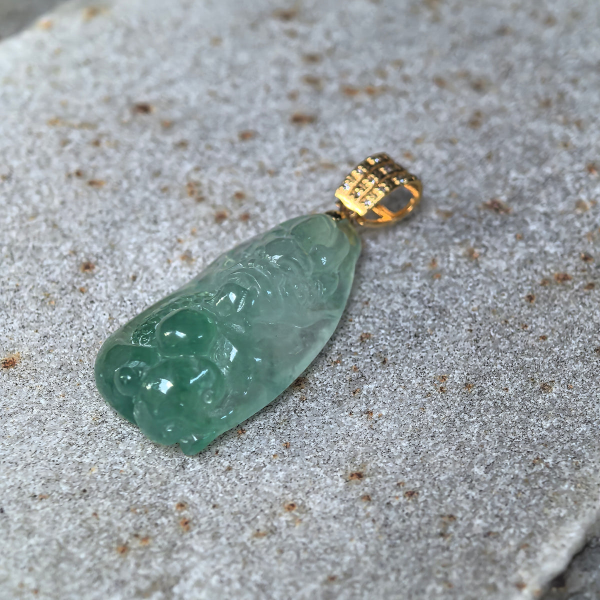 Angled view of a light green jadeite dragon pendant on a stone surface, highlighting the raised carving, detailed scales and gold bail.