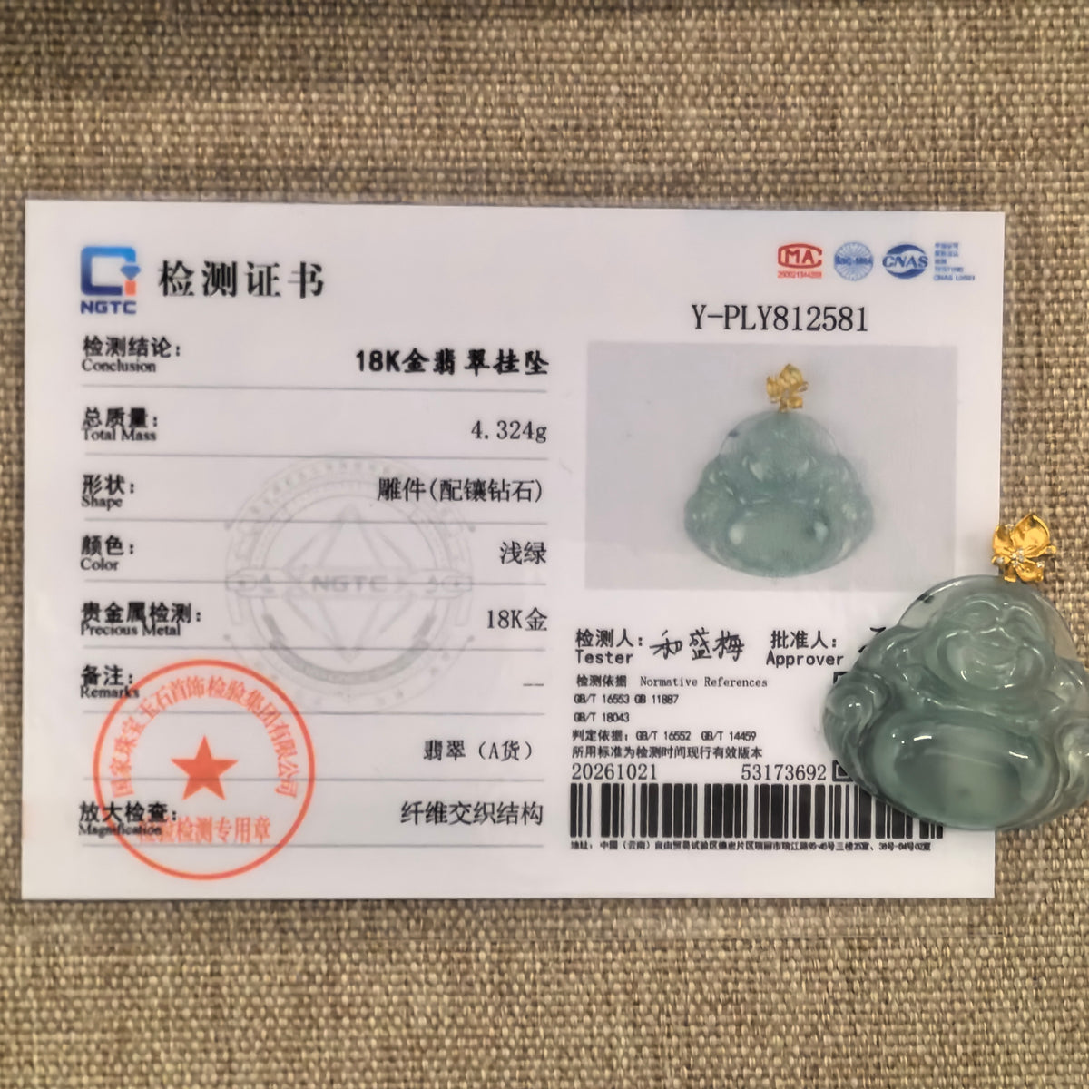 Light green jadeite Laughing Buddha pendant with an 18K gold bail beside its matching NGTC certificate on woven fabric.