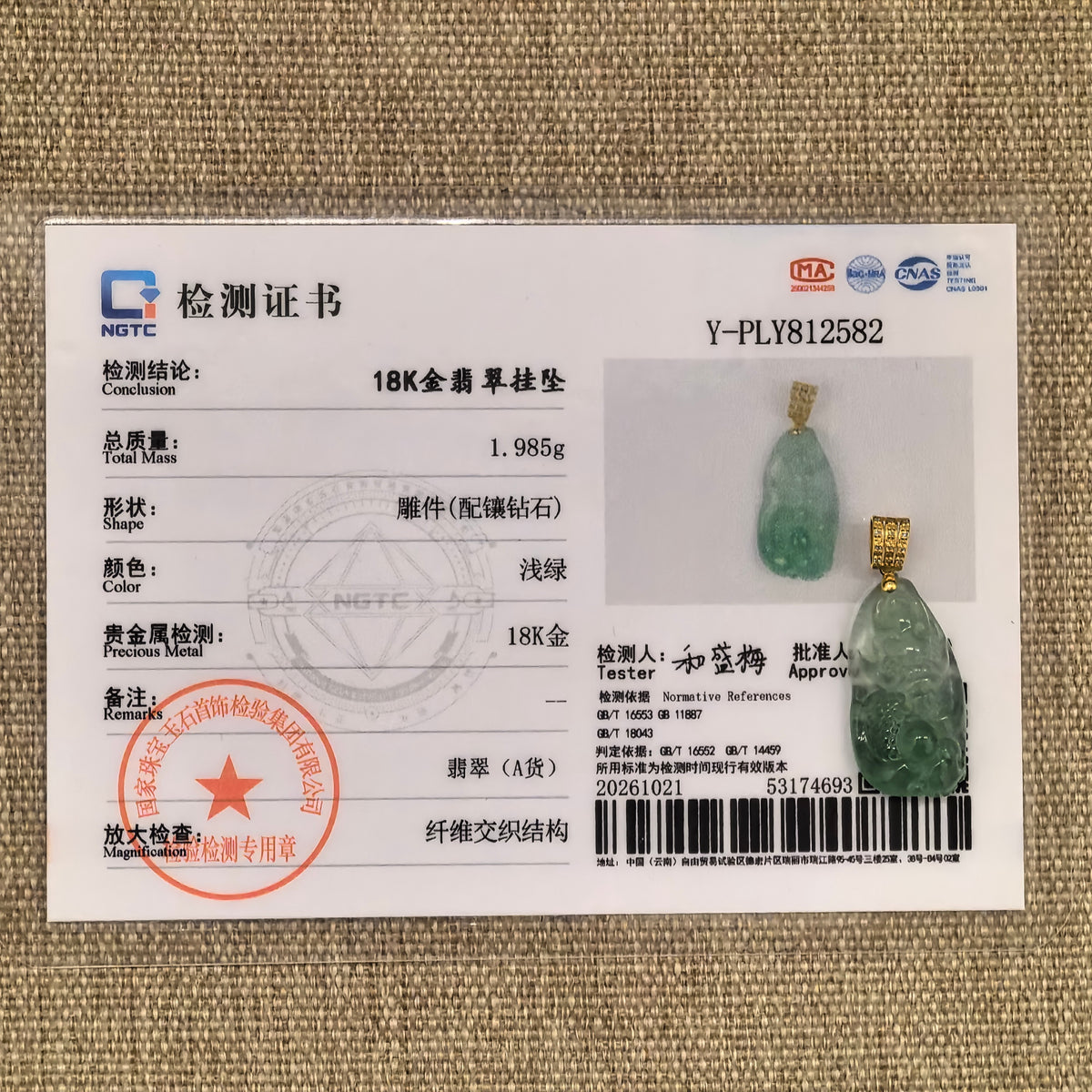 Light green jadeite dragon pendant beside its matching NGTC certificate on woven fabric.