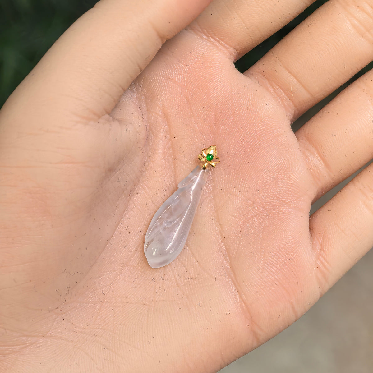 Icy white jadeite Magnolia Pendant resting in a palm, showing its slender petal carving and 18K gold floral bail with a green accent stone.