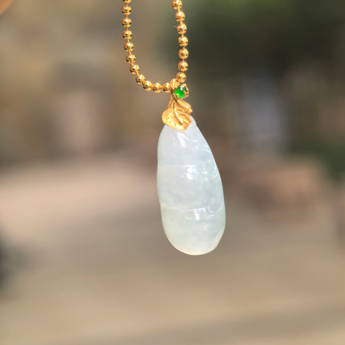Icy glutinous pale green jadeite Bean Pod Pendant hanging from a chain, with an 18K gold leaf-shaped bail and a small green accent stone.