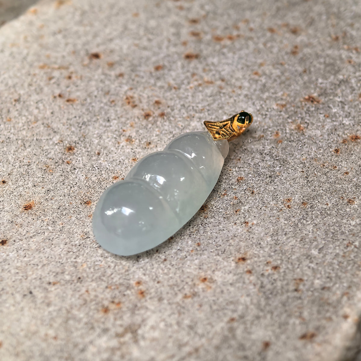 Angled view of a pale green jadeite Bean Pod Pendant on stone, highlighting the domed bean forms and gold leaf detail.