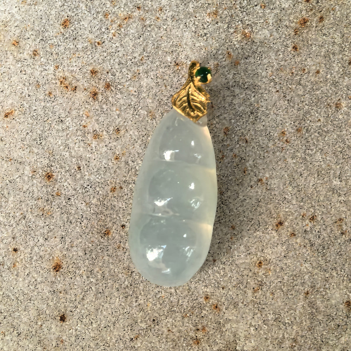 Front close-up of a pale green jadeite Bean Pod Pendant on stone, showing its curved outline, rounded carving and green accent above the gold leaf bail.