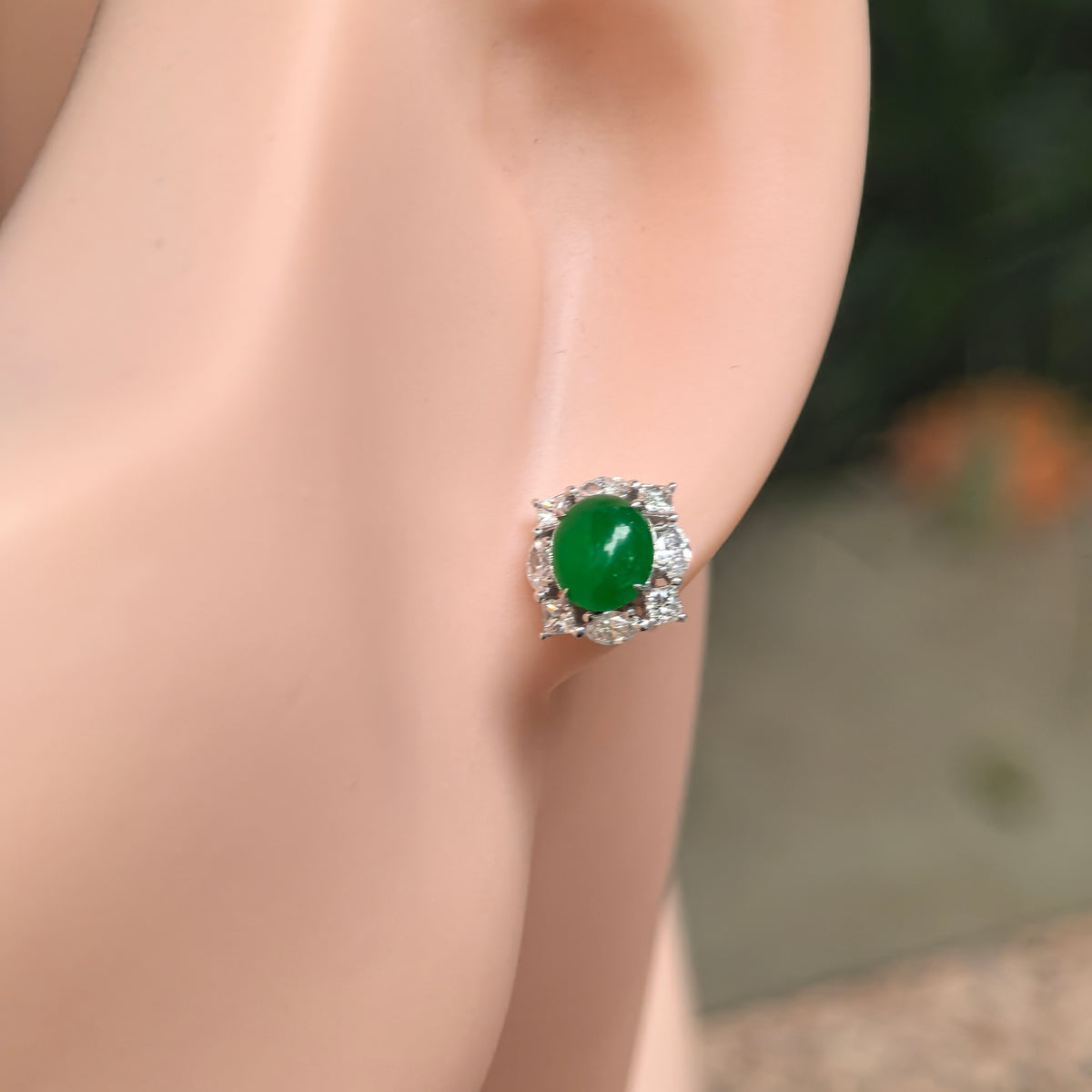 Icy imperial green jadeite stud earring worn on the ear, showing an oval cabochon surrounded by a natural diamond halo in 18K gold.