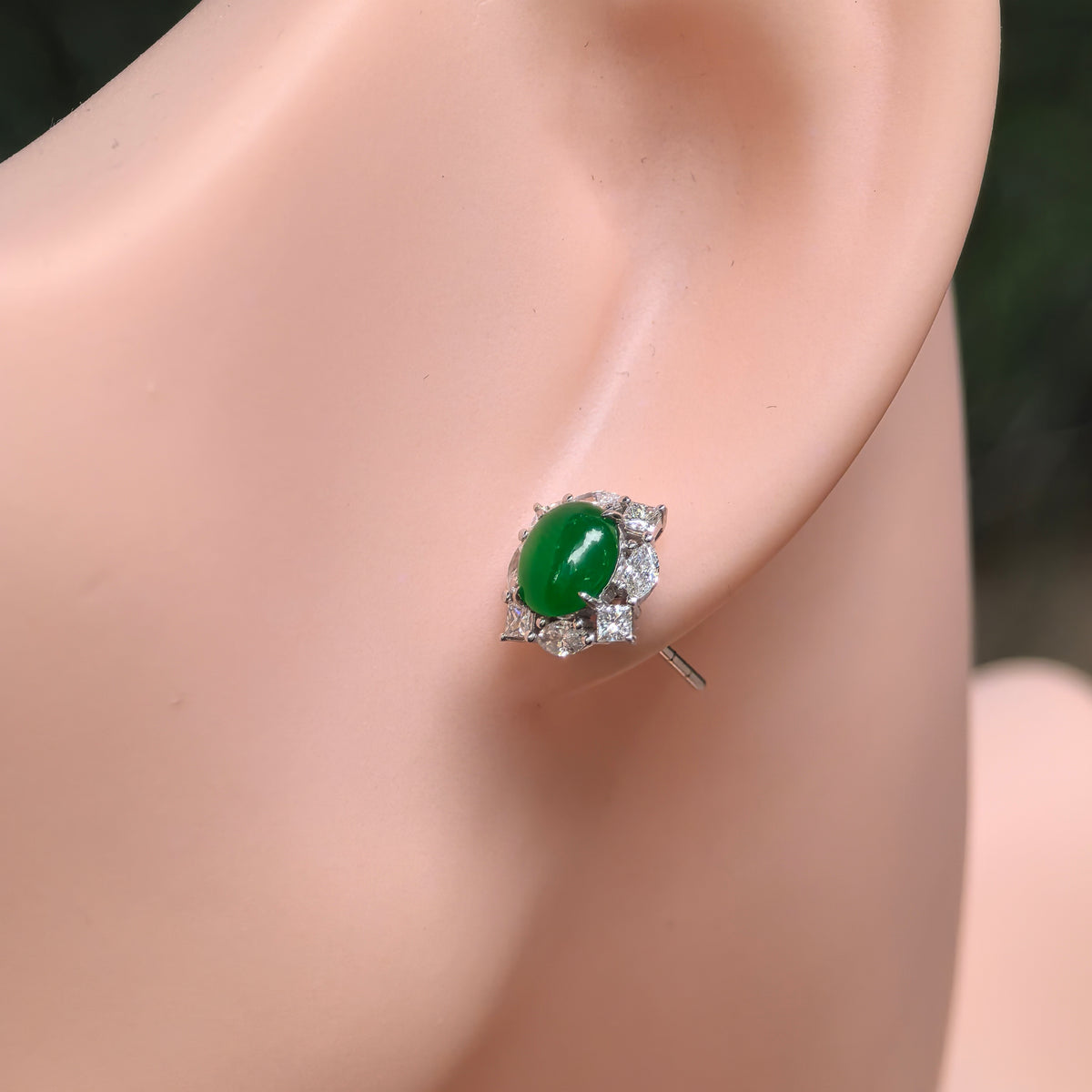 Close-up of an imperial green jadeite stud earring on the ear, showing the polished oval center and alternating square and oval natural diamonds.