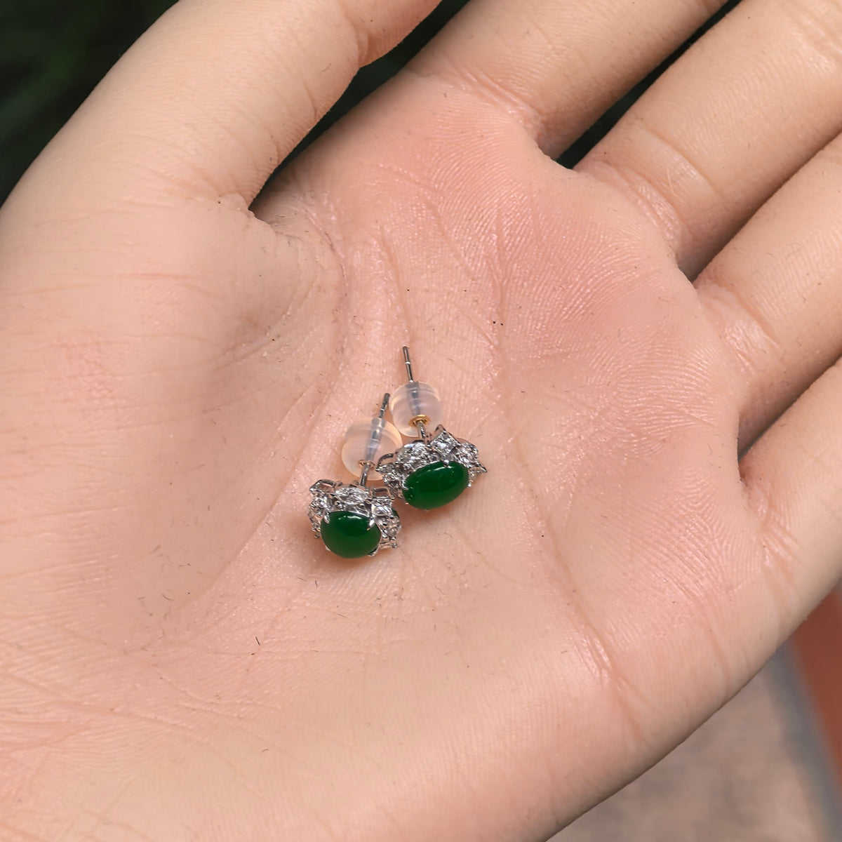 Pair of green jadeite and natural diamond stud earrings in 18K gold resting in a palm, with posts and translucent earring backs visible.