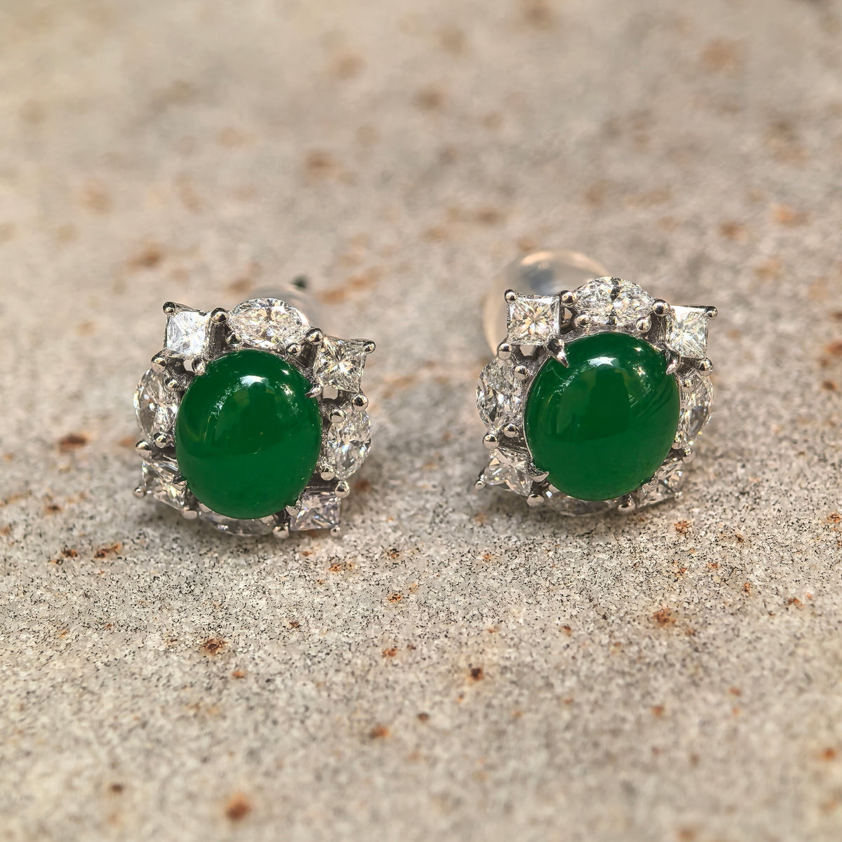 Front view of imperial green jadeite stud earrings on stone, showing oval cabochons framed by square and oval natural diamonds in 18K gold.